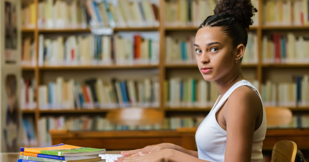 Black female college student sitting at a table in a library, looking out